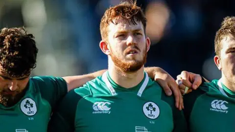 Ciaran Booth Ciaran Booth stands with teammates wearing a green Ireland rugby shirt before a match.