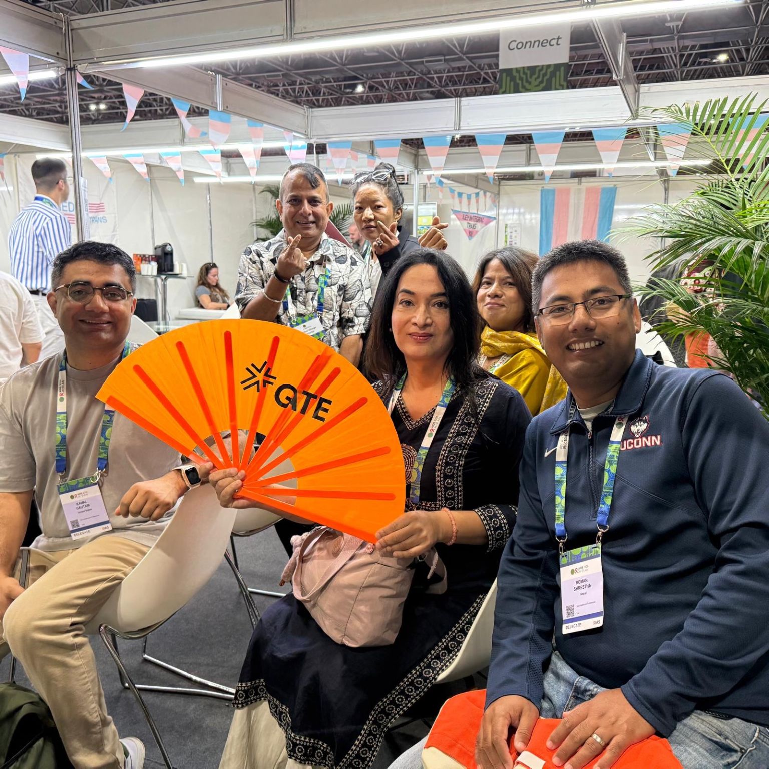 A group of people in a brightly lit conference hall, some sitting, some standing. In the foreground are professors Kamal Gautam and Roman Shrestha and a person holding a bright orange fan. In the background are three people holding up finger heart gestures, and a banner with the transgender flag.