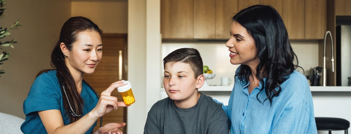 A doctor holds a bottle of pills in her hands while seated next to and talking with a mother and her son