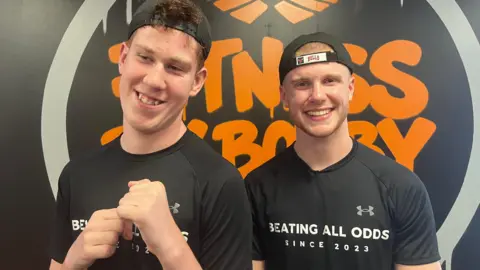 BBC Shay and Bobby are both wearing black T-shirts and caps facing backwards. They are standing in front of a black and orange Fitness By Bobby logo.