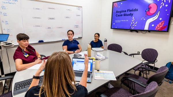 Three nursing students wearing scrubs sit at a conference table listening to an instructor while a screen at the front of the room displays the words "Regina Fields Case Study."