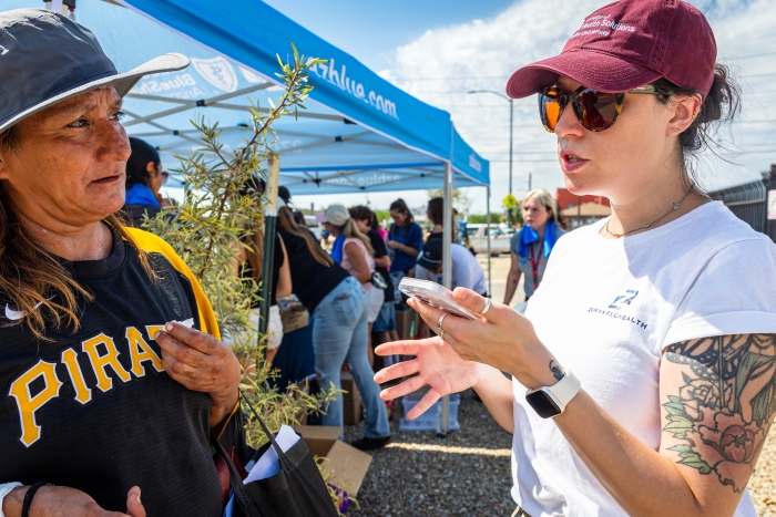 Woman in a white T-shirt, maroon hat and sunglasses talks to a woman in a canvas sunhat while holding a smartphone and standing in a parking lot.