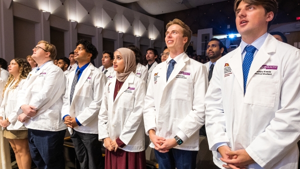 Students in white coats recite an oath in a large auditorium.