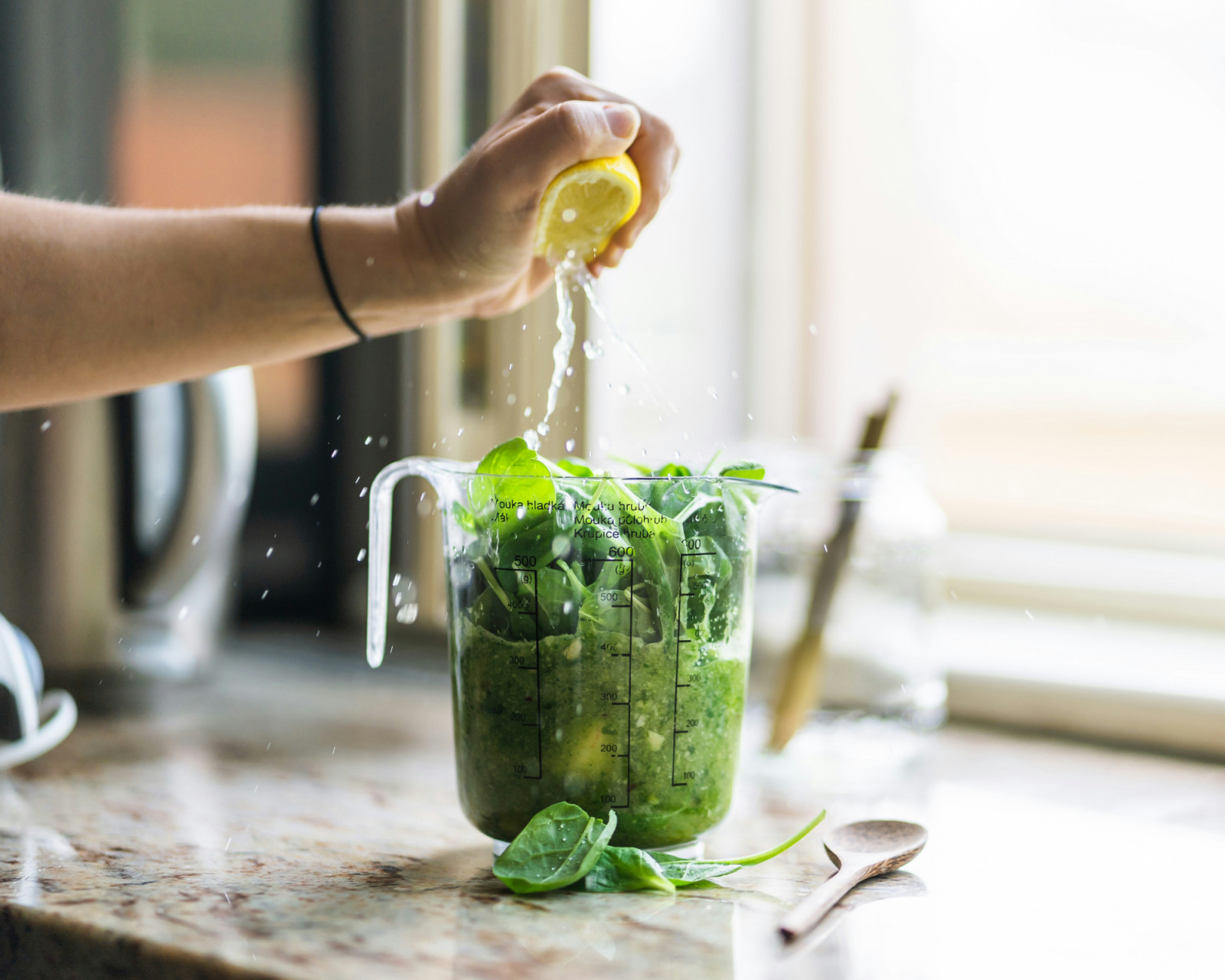 A hand squeezes fresh lemon juice into a measuring cup filled with spinach for a green smoothie.