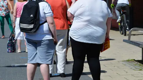 PA Media Two people are standing in the street and their T-shirts are tight over their largely-proportioned bodies. You cannot see their faces, only their backs and legs. There are other people around them and a cyclist to their right.