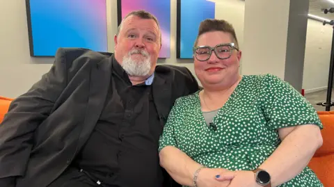 A couple are sitting on an orange sofa. On the left, the man has short dark hair and a gre goatee beard. He is wearing a black shirt and suit jacket. His wife on the right has short hair with an undercut, pattered glasses and a green dress with white spots. They're both smiling.