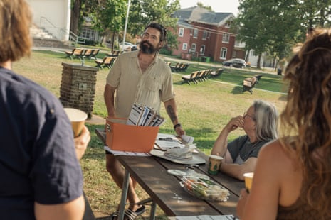 Man stands at end of picnic table in grassy, flat park, with other people sitting and standing.