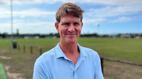 BBC Martin Knight - a man in a blue polo shirt is pictured next to a sports pitch. There is a blue sky behind him. He has cropped brown hair and is smiling.