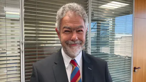 Dr Jason Lane has curly grey hair and is wearing a black suit, white shirt and multi-coloured tie. He has a beard and is smiling. He is standing in an office environment.