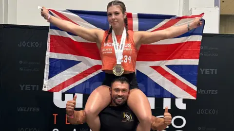 @mayhemathlete Keira, wearing a red sleeveless shirt, black shorts and a large gold medal around her neck, sits on Joel's shoulders as she holds the union flag behind her. Joel, wearing a black shirt, is smiling and has both his arms raised with his thumbs up.