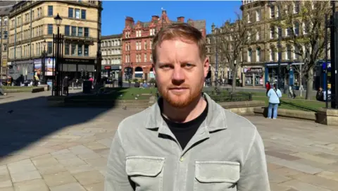 James Grimes is standing in a town with buildings in the background. He has short red hair and a beard. He is wearing a grey shirt over a black T-shirt.