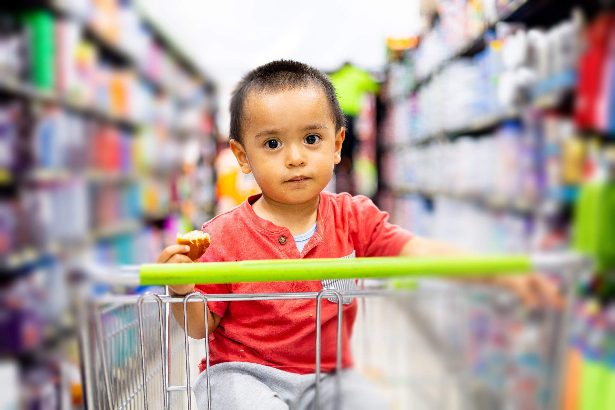 A toddler in a grocery cart holds a bread roll that has been partially eaten.