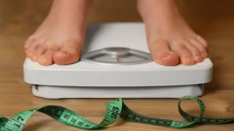 Getty Images Person on scales in bare feet. The scales are white with a red needle and there is a green fabric tape measure on the wooden floor in front of them.
