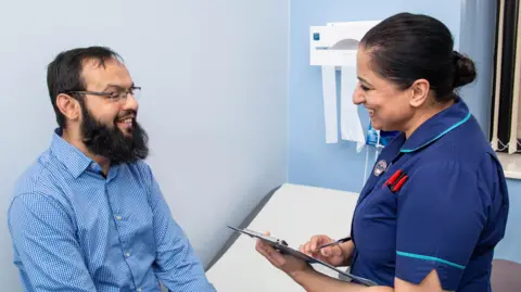 Bradford Teaching Hospitals NHS Trust A man with short black hair and beard, wearing a blue shirt, sits on a hospital bed while a nurse with a clipboard talks to him