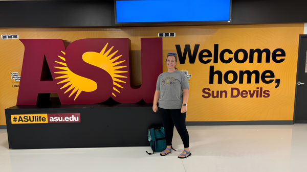 A smiling woman stands in front of the ASU logo. She is wearing an ASU shirt, leggings and sandals. Text on the wall behind her reads: Welcome home, Sun Devils.