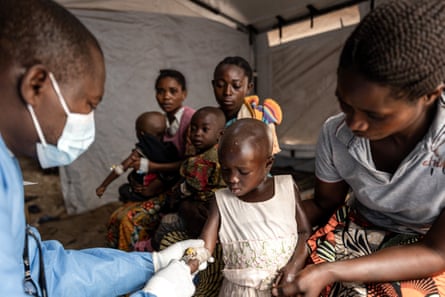 A young girl watches as a masked medic uses a canula in her arm, while two other mothers waiting with their children in the background