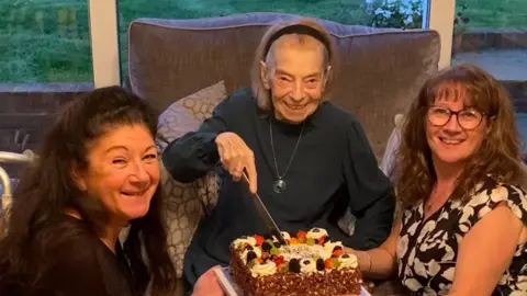 Family handout An elderly woman sat on a sofa cutting into her birthday cake that has lots of fruit on it. She has short hair that has been pushed back with a headband and she is wearing a dark black jumper and a long necklace. She is smiling at the camera. On the right another woman is holding the cake while kneeling next to the elderly woman. She is wearing a sleeveless black top with white flowers and she is smiling at the camera while wearing black frame glasses. She has curly brown hair. Another woman is kneeling on the other side and has black curly hair and is also smiling at the camera.
