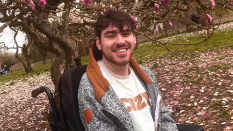 Jimmy Macfarlane/@jimmy.mac01 A smiling young man is seated outdoors in a park beneath blooming trees with pink flowers and fallen petals covering the ground. He has dark wavy hair and a light beard, and is wearing a gray and brown hoodie over a white shirt that says cozy, the backrest of his wheelchair is visible behind him.
