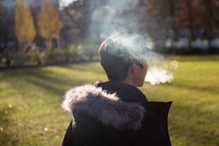 A young person with short dark hair and wearing a jacket with a furry hood, seen from behind, exhales vape smoke around them.