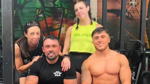 @mayhemathlete Two men, one shirtless and one wearing a black shirt, smile as they sit down in a gym. A woman has her arms on the shoulders of a man on the left as she crouches down slightly. A girl wearing a yellow shirt and black shorts stands behind them.