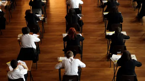 PA Rows of pupils doing exams in a hall. Some are wearing blazers while others have white shirts on. There is a mix of girls and boys and all have their heads down and are facing their work so we can only see the backs of their heads.