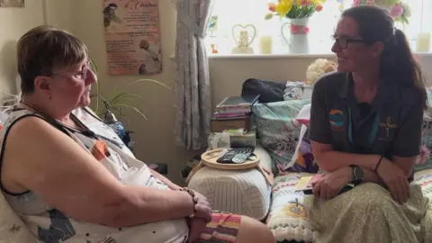 The image shows two people seated facing each other in a living room, appearing to be having a conversation. One person is sitting on a sofa covered with patterned cushions and a floral throw, while the other is seated nearby wearing a grey polo shirt and light-coloured skirt, with both positioned beside a large window.