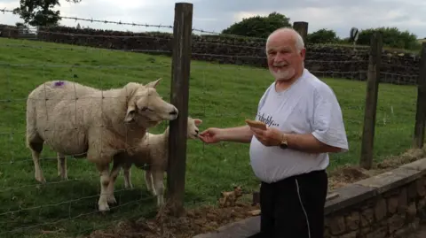 Mick Wadge Mick Wadge - an older man with receding white hair and a white beard and moustache - stands beside a field, feeding two sheep through a wire fence