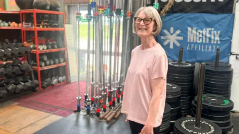 Krish Stevens An older woman wearing a pink T-Shirt with glasses standing in front of gym equipment