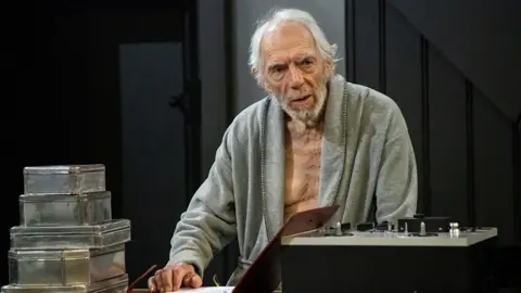Stagefright Films Peter Marinker in dress rehearsals for the play Krapp's Last Tape. He is wearing an old grey dressing gown and sitting in front of a desk. There is a tape recorder in front of him and some silver boxes containing reel-to-reel tapes.
