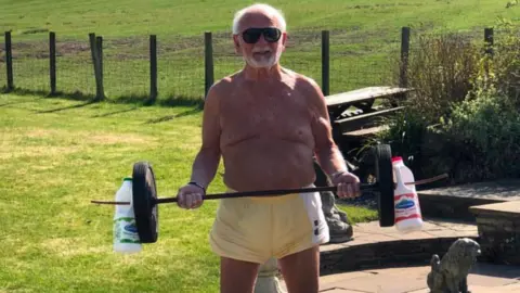 Mick Wadge Mick Wadge stands on a patio holding a homemade barbell weighted with wheels and milk containers