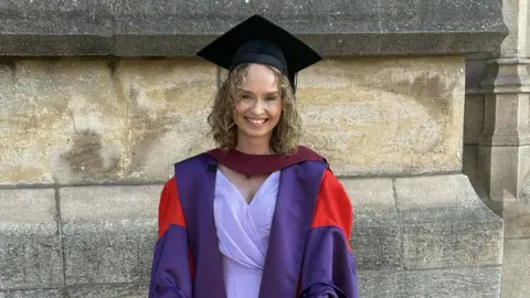 Family photo A woman in a graduation gown smiles at the camera. She is wearing a black mortarboard and a purple and red graduation robe. Beneath that she is wearing a lilac outfit. She has curly dark blonde hair. Behind her is a stone wall.