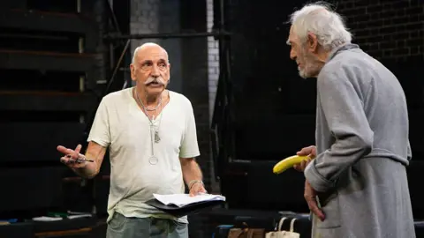 Stagefright Films Dave Wybrow, with white hair and a handlebar moustache, is wearing a white t-shirt and talking, on stage, to Peter Marinker who is wearing a grey dressing gown and holding a banana as a prop.