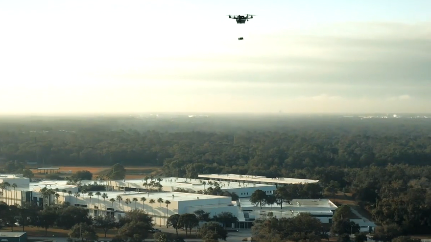 wide shot into the sky of a drone hovering and dropping down a supply box on a tether toward the ground, where there is a large building