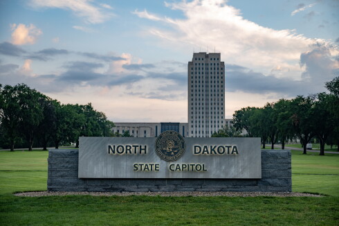 A gray stone sign reads North Dakota State Capitol in the foreground with trees, a grassy field and a tall gray building in the background. The letters on the sign are backlit and the signs bears the seal of North Dakota.