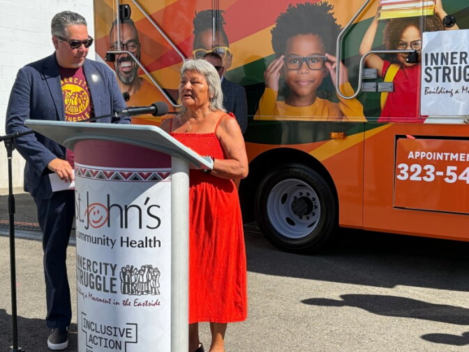 An older woman with medium skin tone, wearing an orange dress, speaks behind a podium next to a man with medium skin tone, wearing a blazer over a t-shirt. They stand in front of an orange bus with photos on the side of it.