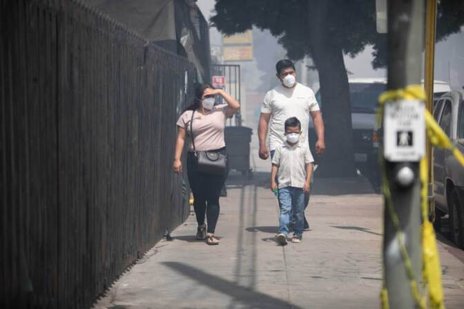 A man, woman, and child, all wearing face masks, walk down a sidewalk past a metal fence with the air filled with smoke.