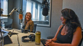 Low-angle photograph of two women sitting at a table in front of microphones inside a professional radio studio. Headphones, a cellphone, a water bottle and an energy drink are visible on the table.