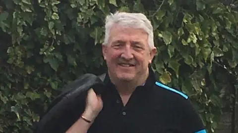NHS England Paul stands outdoors for a photo. He has grey hair and is smiling at the camera