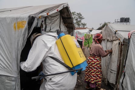 A person in a hazmat suit enters a tent with a yellow cylinder on their back while a woman in a colourful dress stands in the background.