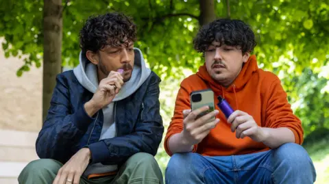 Getty Images Two young men sit outside vaping. They are chatting, while looking at a mobile phone.