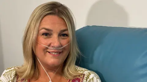 BBC A picture of Helen on a chair in the waiting area of the lung function test area at Nottingham's Queen's Medical Centre. She is smiling at the camera and on display are tubes on her face into her nose which are connected to a portable oxygen machine