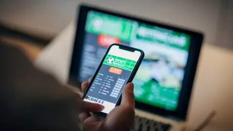 Getty Creative/iStockphoto A phone is being held in front of a blurry person and it shows a gambling website. There is a laptop showing a blurry gambling screen in the background.