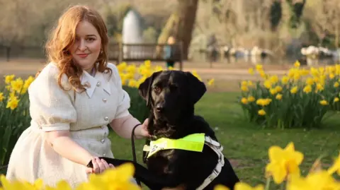 Emily Davison/fashioneyesta2012 Emily, a young woman with mid length auburn hair, kneels in a park beside Rosie, a black Labrador guide dog, gently holding her paw. Emily is wearing a light cream dress and sitting among bright yellow daffodils, with flowers in front of her and behind her. Behind them is a large tree trunk, a fountain, and out of focus greenery.