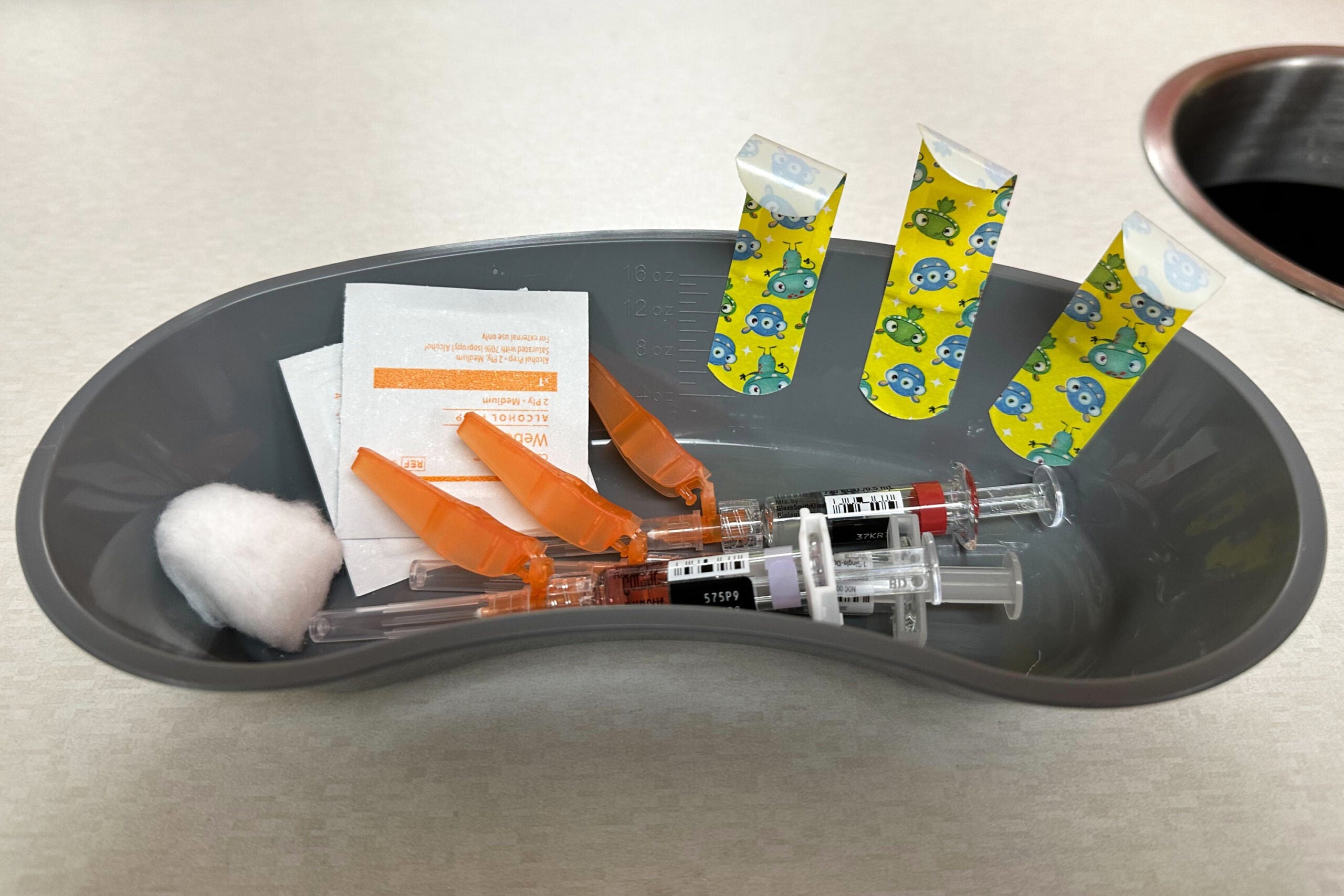 A medical tray holding two syringes, alcohol wipes, a cotton ball, and three cartoon bandages with monsters printed on them.