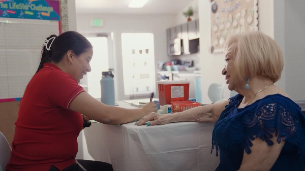 A health care worker in a red shirt prepares a blood pressure monitor for an elderly woman in a navy-blue lace top. They are in a brightly lit room with medical supplies on a table.
