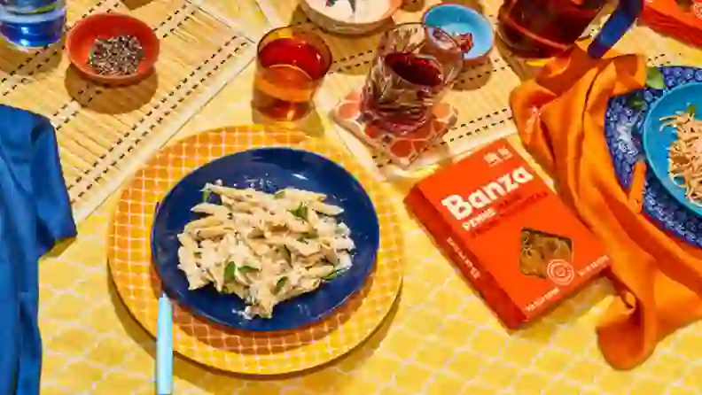 Box of Banza chickpea penne beside a plated pasta dish and drinks