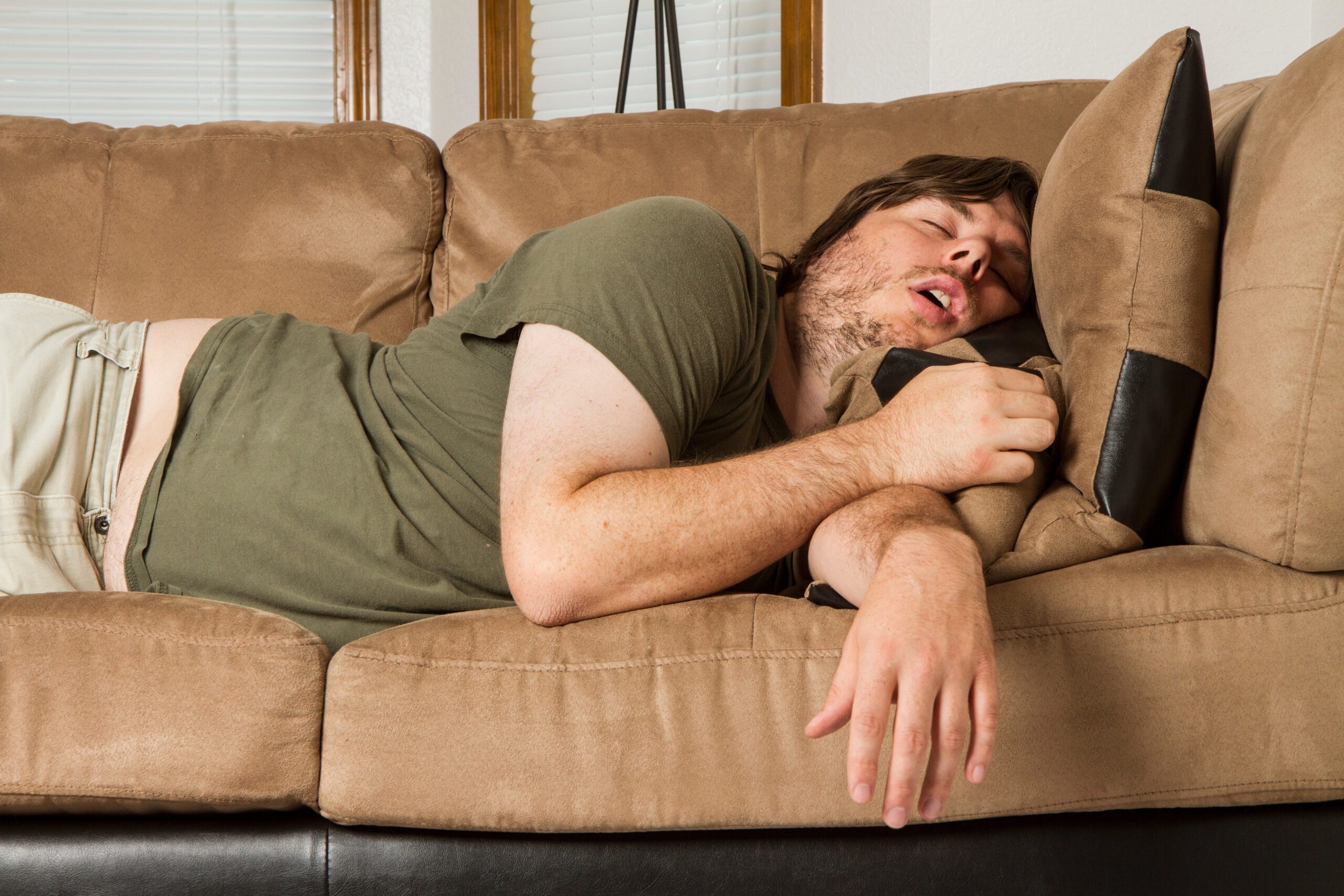 Person asleep on a couch, wearing casual clothes, mouth open, holding a pillow. The setting suggests relaxation or fatigue
