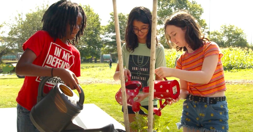 Three Kids Watering Young Plants With Watering Cans In An Outdoor Garden Setting On A Sunny Day