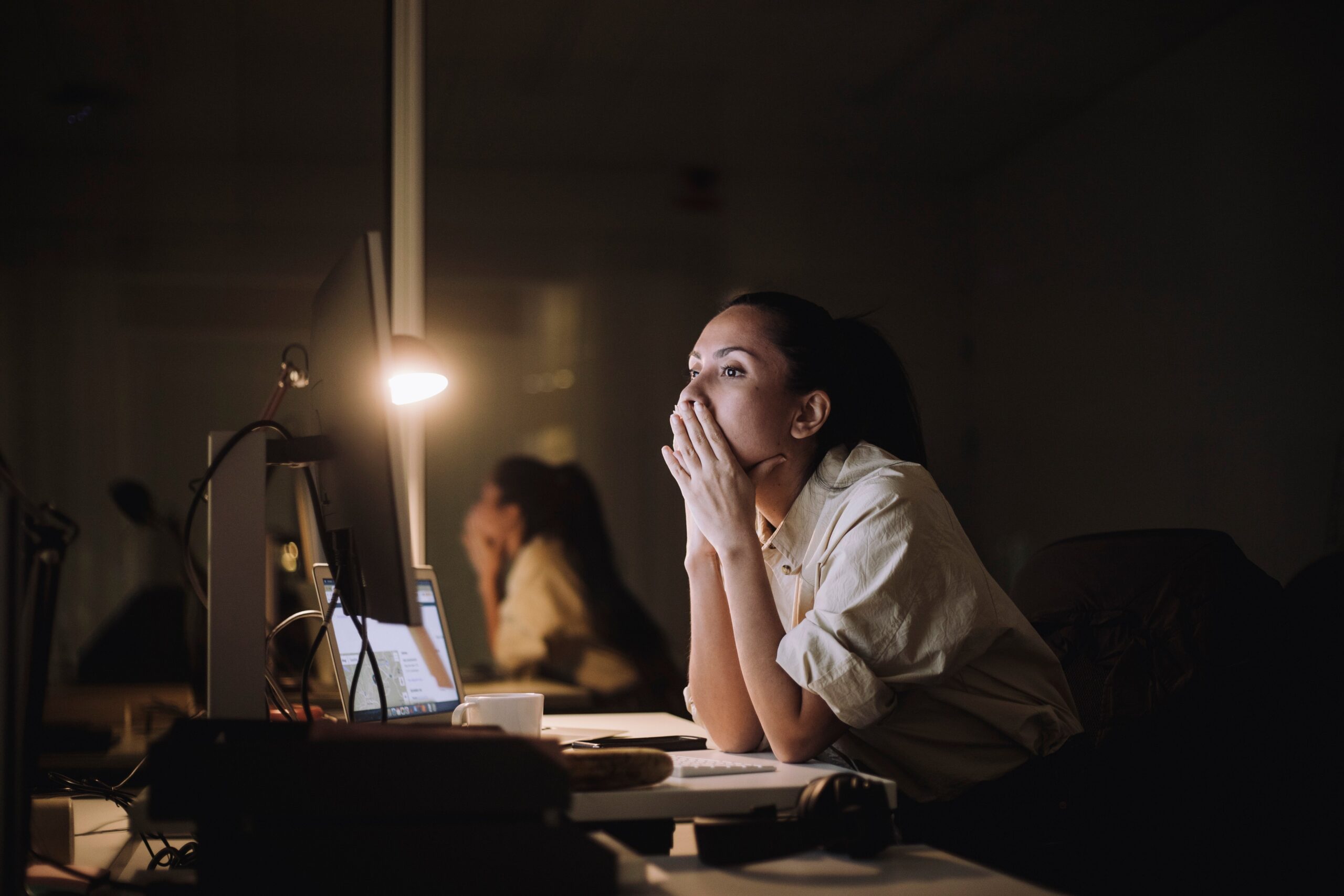 Person in a dimly lit office, looking thoughtfully at a computer screen with hands on face, suggesting deep concentration or contemplation