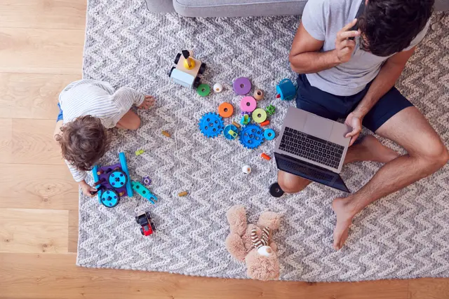 Overhead image of a dad on his laptop and talking on the phone, sitting on floor next to a child playing with toys and blocks